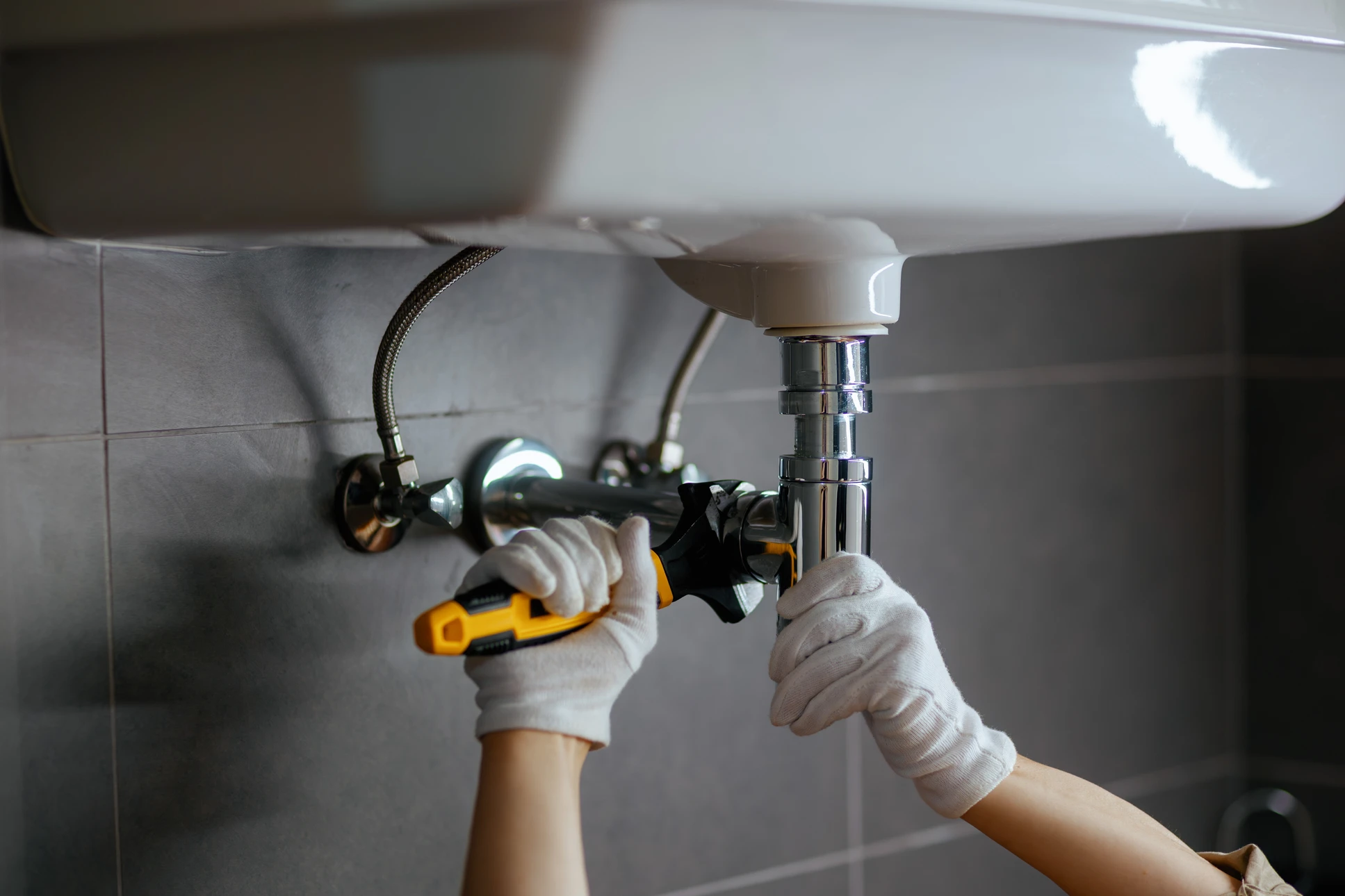 Photographe réaliste d'un plombier professionnel au travail sur un chantier de rénovation de salle de bain. Le professionnel, vêtu d'une combinaison de travail, intervient activement sur l'installation de tuyauterie ou d'équipements sanitaires. L'arrière-plan montre l'environnement réel d'un chantier intérieur avec éléments de rénovation visibles. La composition capture l'expertise et le professionnalisme du métier de plombier qualifié.